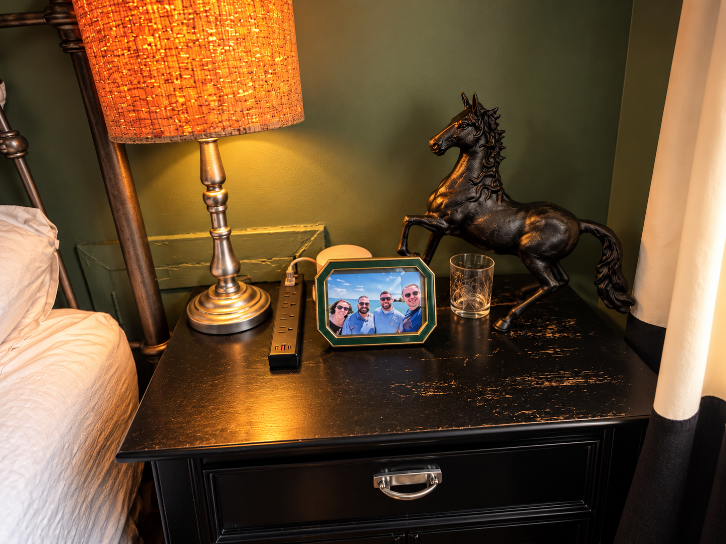 Bedside table with warm lamp, framed personal photo, drinking glass, and black horse sculpture