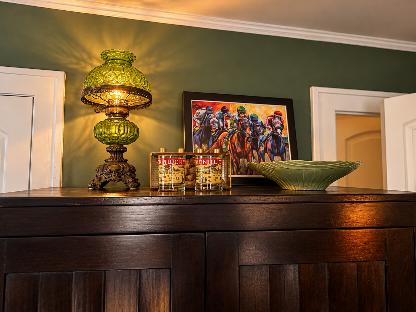 Dark wood cabinet topped with a green antique lamp, Kentucky keepsakes, horse artwork, and a green bowl