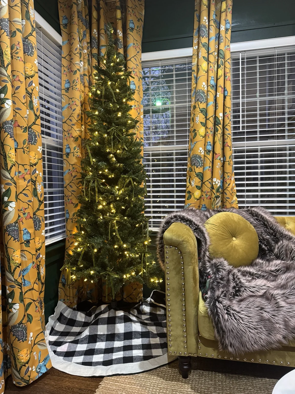 Library at Christmas with small lit tree beside mustard floral curtains and velvet sofa