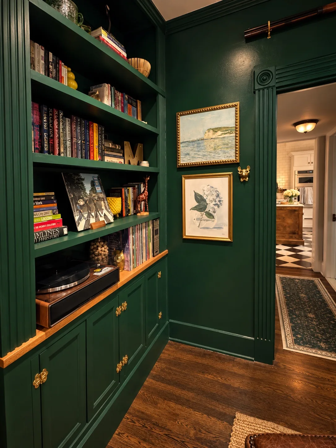 Detailed view of custom deep-green library built-ins with books, records, brass hardware, art, and view toward the kitchen