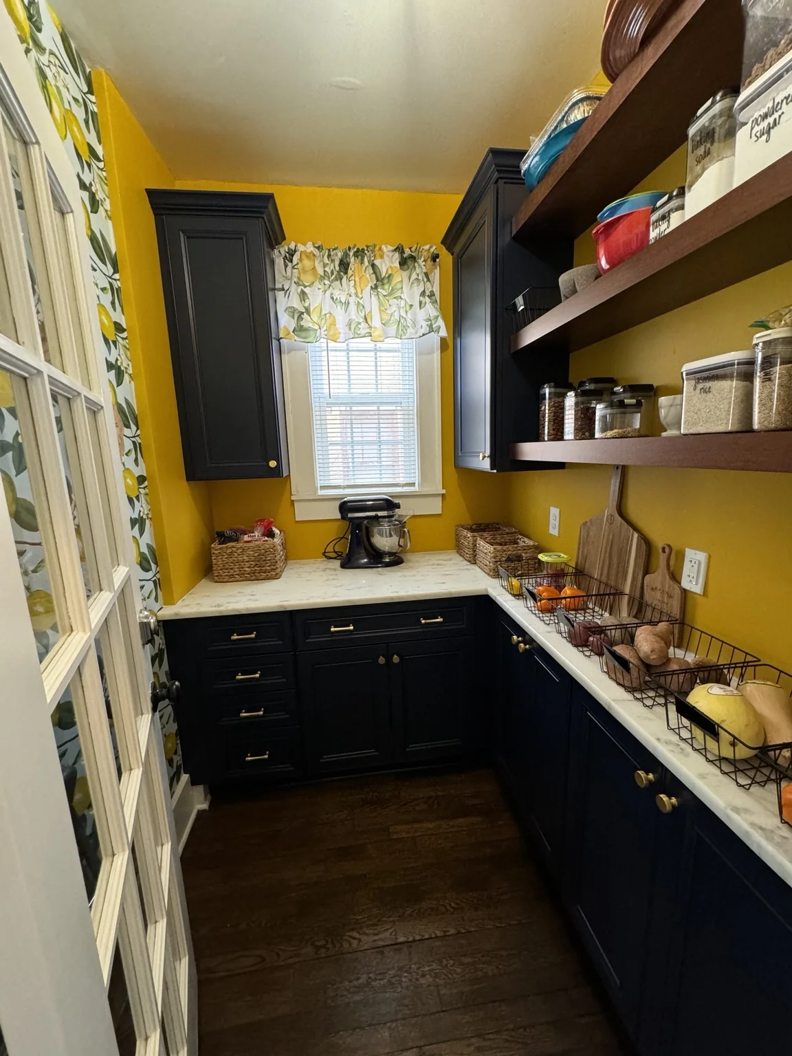Finished pantry with bold yellow walls, dark blue cabinetry, brass hardware, warm wood shelving, white counters, and lemon details